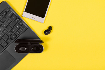 Laptop, smartphone, charging case and wireless earpieces on yellow background with copy space