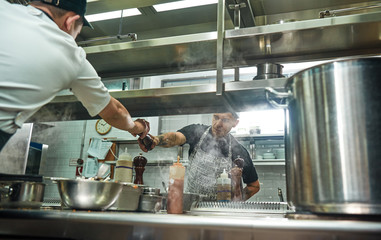 Take it. Young professional chef in apron giving a pepper grinder to his assistant while cooking process