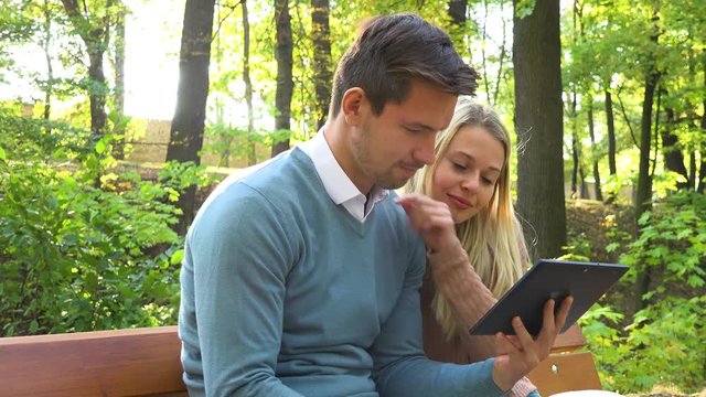 A young attractive couple looks at a tablet on a bench in a park on a sunny day - closeup