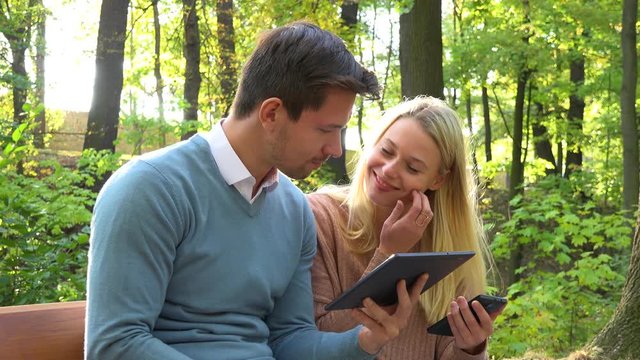 A young attractive couple works on a smartphone and a tablet on a bench in a park on a sunny day - closeup