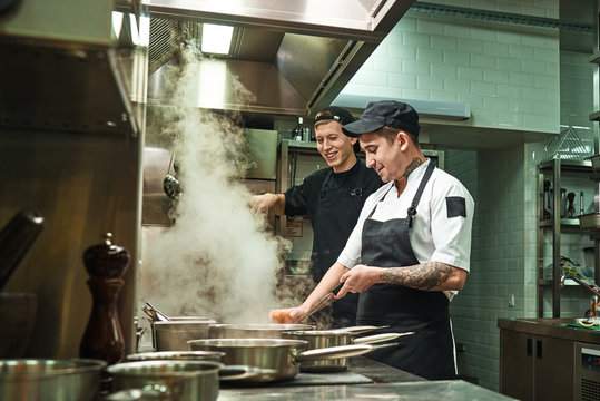 Positive mood. Side view of two cheerful and smiling cooks in uniform are preparing a food in a kitchen restaurant