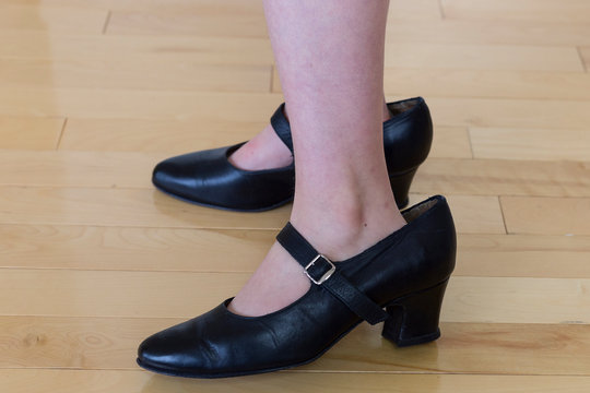 Child Standing Wearing Adult Black High Heeled Shoes Too Big For Her, Against Hardwood Floor