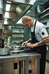 Last details. Side view of confident male chef in black apron garnishing his dish in a restaurant kitchen.