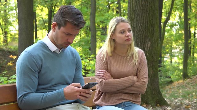 A man and a woman sit on a bench in a park, the man is absorbed with his smartphone, the woman is angry at him