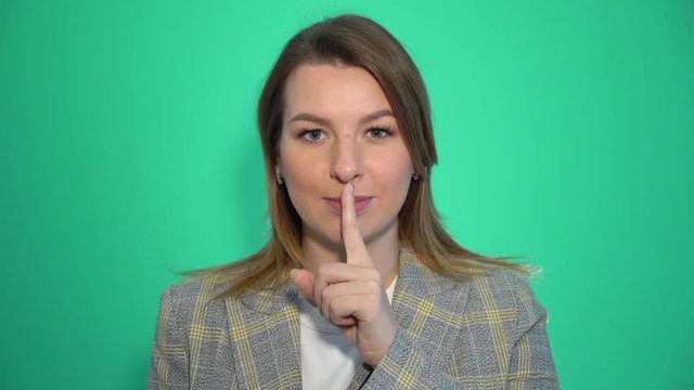 Silence, please! Close up portrait of young woman holding her forefinger on lips showing hush silence sign, standing over green background chromakey