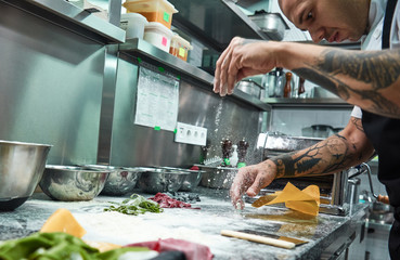 Making pasta process. Close up photo of concentrated chef with black tattoos on his arms pouring flour on kitchen table while making pasta