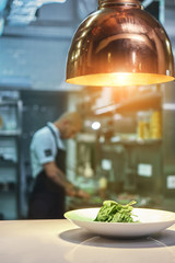 Ready to serve. Vertical photo of fresh green salad standing on the table under the light with working chef on the blurred background.