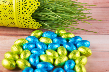 Green grass stems laying in flower pot on wooden table naer variety of small dessert eggs in foil. Selective focus