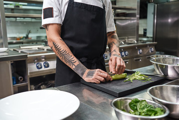 Simple task. Close up of male hands with beautiful tattoos peeling cucumber for salad while standing in a restaurant kitchen