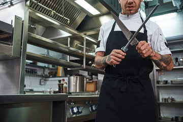 Professional chef. Brutal young man in apron with several tattoos on his arms sharpening a knife in...