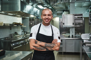 I love my work Cheerful young chef in apron keeping tattooed arms crossed and smiling while standing in a restaurant kitchen - Powered by Adobe