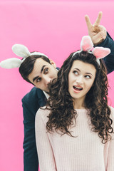 Young couple on pink background. On the head is a rabbit ears. The man shows two fingers at the top of his wife's head. The girl opened her mouth with amazement. Easter.