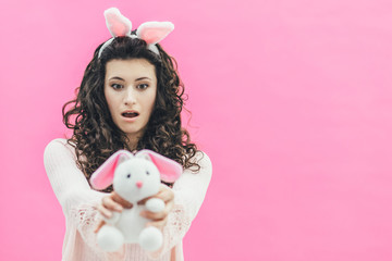 Young pretty girl standing on a pink background. On the head of the ears of the bunny. During this, he holds a soft toy in a bunny, isolated on the background of the girl. Easter.