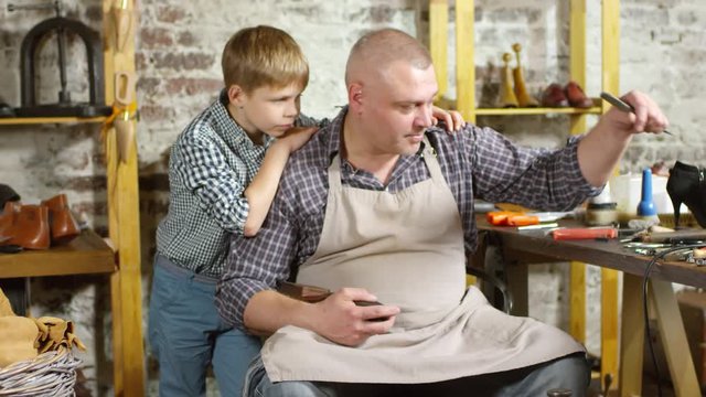 Zoom in of middle aged shoemaker repairing shoe sole and explaining process to little son standing beside him and embracing