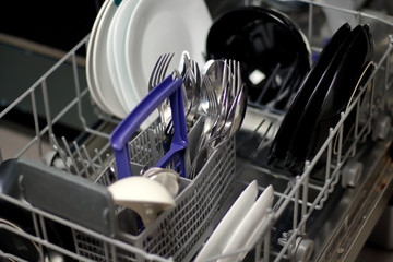 Dishwasher with white and black plates with bowls, forks, spoons, knives in a stand basket, clean washed dishes in a dishwasher