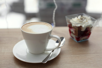 White cup of hot cappuccino on white saucer and red velvet dessert on wooden bar table next to window. Cold snowy weather outside