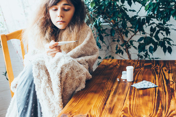 Girl wrapped in a blanket looking at the thermometer. Sick woman with flu.
