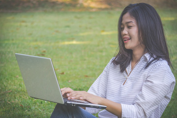 The asia woman is playing computer on a notebook.She sits and play laptop in park.