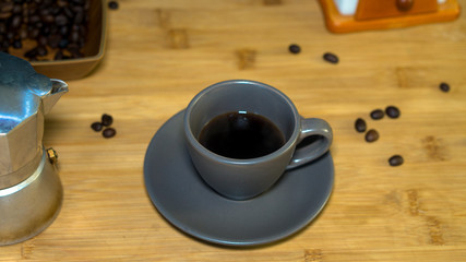 A cup of espresso coffee, put on a wooden table, an Italian coffee maker (moka) and in the background roasted coffee beans in a pot