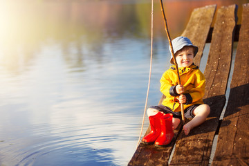 Little boy siting on wooden dock and fishing at sunset.