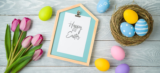 Easter holiday background with easter eggs in bird nest, note and tulip flowers on wooden table.