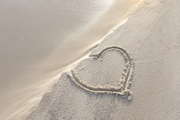Fototapeta na wymiar Heart shape written in sand on the beach with warm sunlight in the morning. High tide and waves come closer. Love in danger, relationship in difficulties and sadness concept.