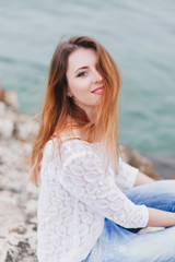 Beautiful young red-haired woman in white lace blouse and blue jeans sitting on a rocky shore near sea and smile. Close up mood portrait.