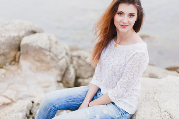 Beautiful young woman in white lace blouse and blue boyfriend jeans sitting on a rocky shore near sea.