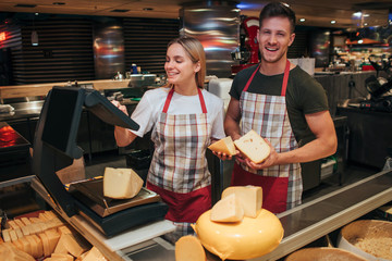 Young man and woman stand at cheese shelf in grocery store. Female model use weights for piece of...