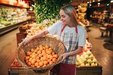 Young woman stand at fruit boxes in grocery store. She hold basket with oranges and look at them. Positive happy worker in store.