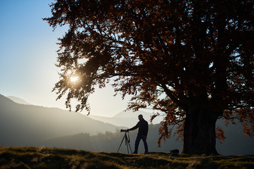 Male tourist photographer with tripod and professional camera standing under large tree with golden leaves, taking picture of woody foggy mountains landscape and bright sunset in blue sky background.