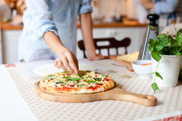 Obraz na płótnie Canvas Woman is cooking pizza in cozy home kitchen. Girl is decorating italian dinner with greens, fresh basil, arugula. Homemade pizza is served on wooden board on table. Lifestyle moment. Close up.