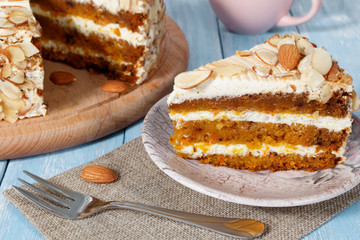 Close-up the slice  of vegetarian carrot cake with almonds on a saucer, a fork and wooden board with cake near on blue wooden table. Pink cup in the background.