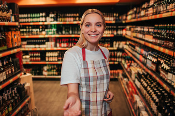 Young woman stand in alcohol shelfs in grocery store. She reaxh hand to camera and smile. Cheerful happy young woman.