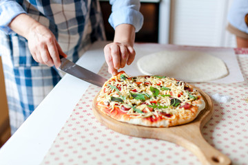 Obraz na płótnie Canvas Woman cuts hot pizza on wooden board in cozy home kitchen. Cooking process of italian family dinner at home. Female hands hold knife and slice of pizza. Lifestyle moment.