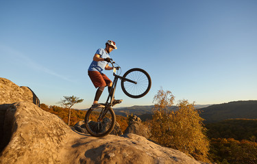 Fototapeta na wymiar Professional cyclist riding on back wheel on trial bicycle. Sportsman biker balancing on the edge of big boulder on the top of mountain at sunset. Concept of extreme sport active lifestyle
