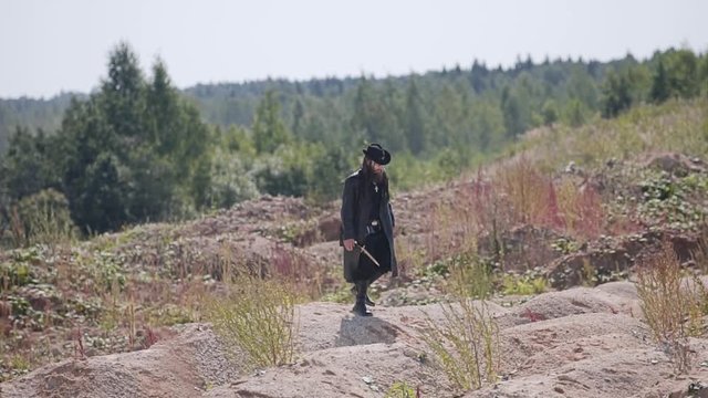 A mysterious man in a black cloak and hat is walking through the desert. Hilly dry land. NN looks around. Behind the background you can see the forest.