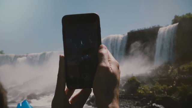 Slow motion close-up shot, human hands holding black smartphone, taking photos of epic Niagara Falls waterfall scenery.