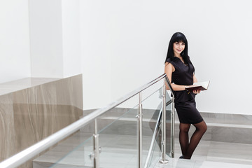 Young Beautiful happy brunette woman dressed in a black business suit working with a notebook, standing in  office, smiling.