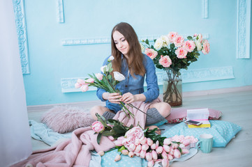 Cheerful young woman sitting on the floor, making flower bouquet of pink tulips in light sunlit room with blue walls.