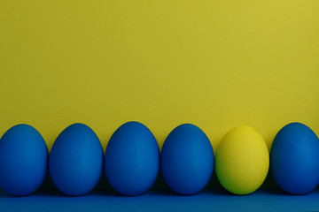 Five blue and one yellow painted Easter eggs stand in a row on a yellow with blue background. Happy Easter holiday card or banner. Copy space.
