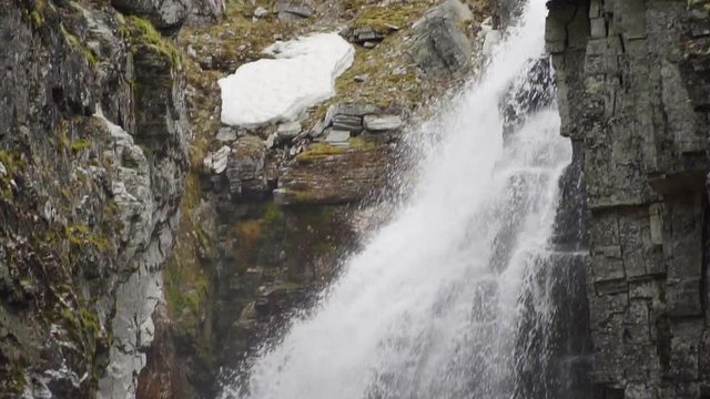 Tilt up at Jutulhogget Waterfall at the Rondane Nationalpark in the Norwegian mountains in slow motion floating down in a big creek of rough granit stone covered in moss, fern, ice and snow.