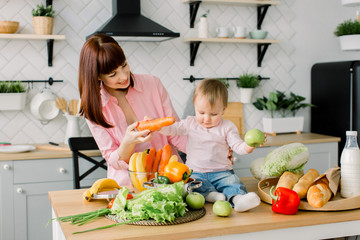 Happy mother showing fresh vegetables to her little baby girl sitting on the wooden table at home kitchen. Family, food, healthy eating, cooking and people concept
