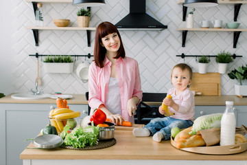 Young pretty mother in rose shirt preparing fresh breakfast and chopping carrot at home kitchen. Little baby girl holding banana and sitting on the wooden table with fresh food, milk and bread