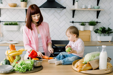 Smiling young mother chopping carrot while preparing fresh salad and little cute baby toddler holding green apple and sitting on the table in kitchen. Baby helps mother to cook at the home kitchen