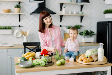 Young mother preparing fresh salad with her little baby daughter and and having fun while putting green oak lettuce in glass bowl in kitchen. Mother and little baby watching at camera at the kitchen