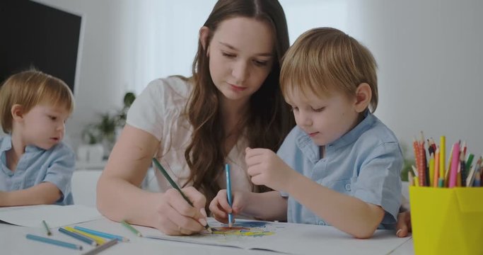 A young mother with two children sitting at a white table draws colored pencils on paper helping to do homework