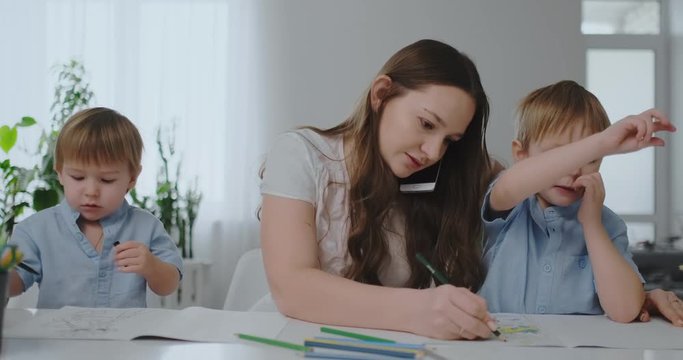 A young mother with two children talking on a mobile phone draws with a pencil and helps children draw with colored pencils