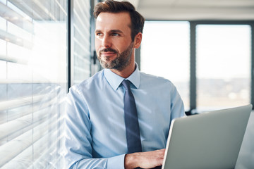 Handsome businessman standing with laptop, looking throught the window in modern office