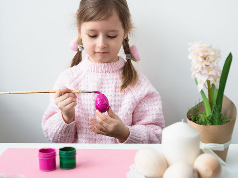 A beautiful little girl with pink bunny ears (hare) is preparing for the celebration of Easter, painting a wooden egg in pink colore. Light background. Free space for text.